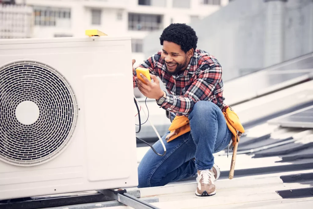 A smiling HVAC technician using a digital multimeter to diagnose an outdoor air conditioning unit on a Boulder rooftop.
