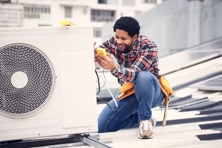 A smiling HVAC technician using a digital multimeter to diagnose an outdoor air conditioning unit on a Boulder rooftop.