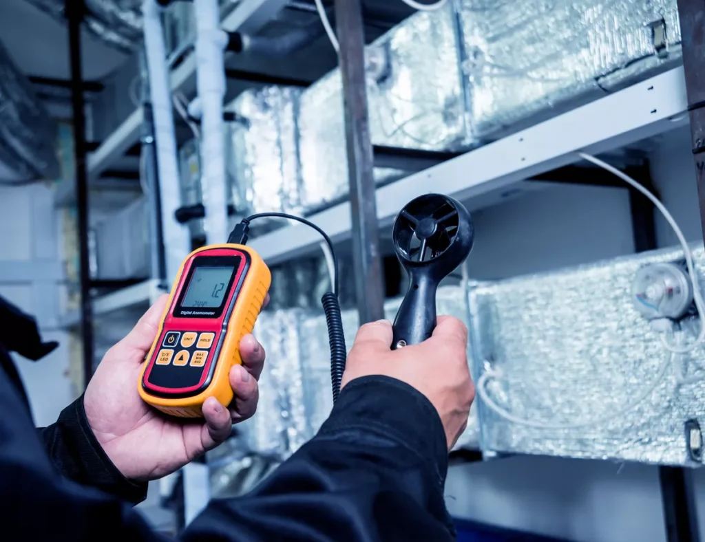 A technician using a digital handheld anemometer to measure airflow and velocity from insulated HVAC ductwork for an energy efficiency audit.