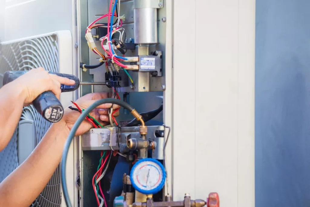 Close-up of a technician's hands using a screwdriver to repair internal wiring and electrical components of an air conditioning unit next to pressure gauges.