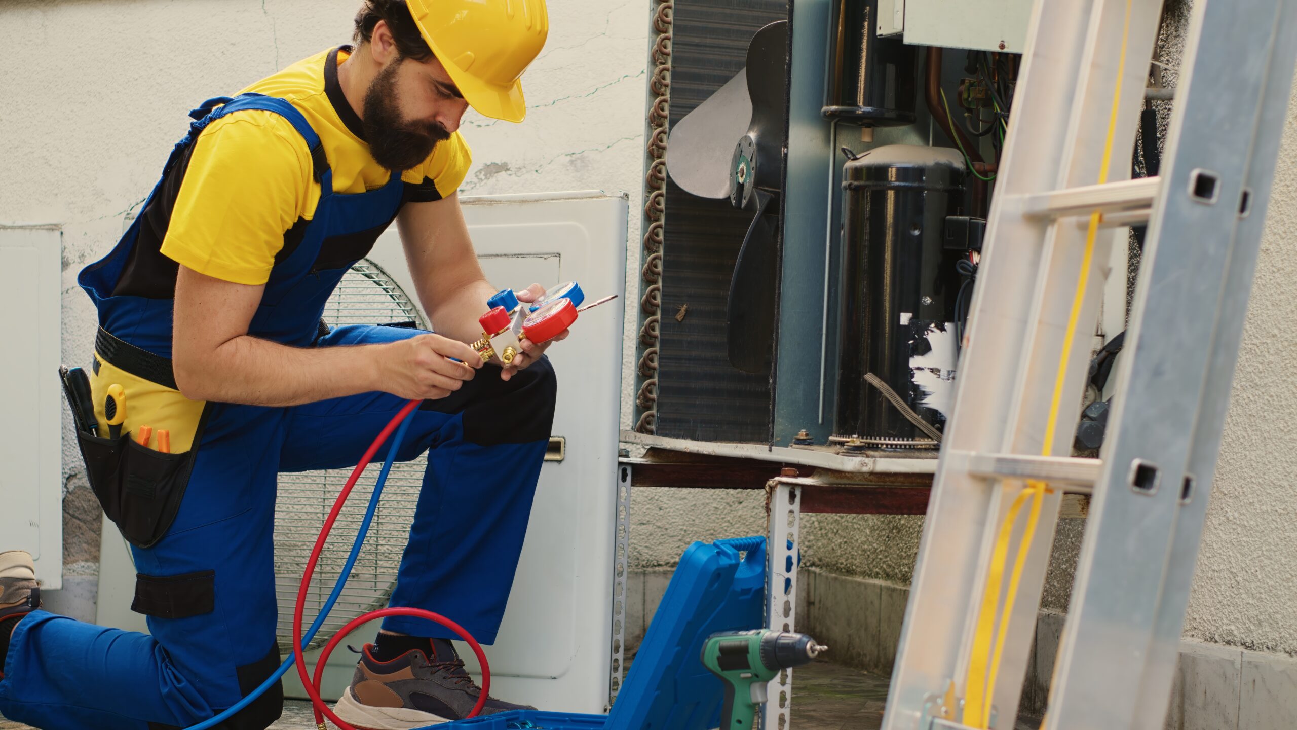 A bearded HVAC technician in a safety hardhat and overalls repairing an outdoor condenser unit with specialized pressure hoses and professional tools.