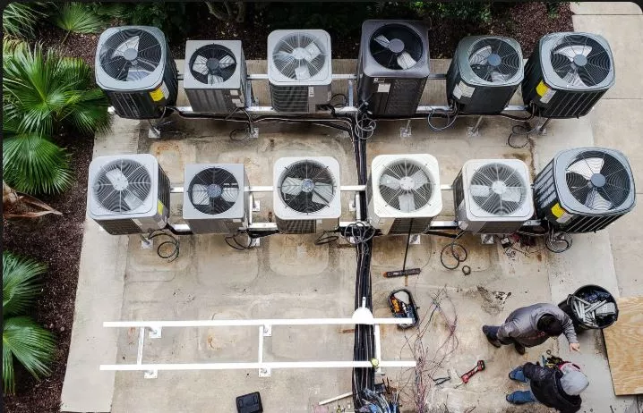 Two professional HVAC technicians performing maintenance and installation on a large array of outdoor air conditioning condenser units on a commercial concrete rooftop.