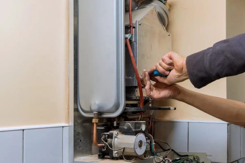 Close-up of a technician's hands using a blue screwdriver to replace components on a water heater in a gray-tiled utility room.